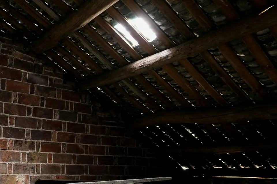 Een ongeïsoleerde zolder: daglicht valt tussen de dakpannen door, boven houten balken, een gemetselde muur en een oude plankenvloer